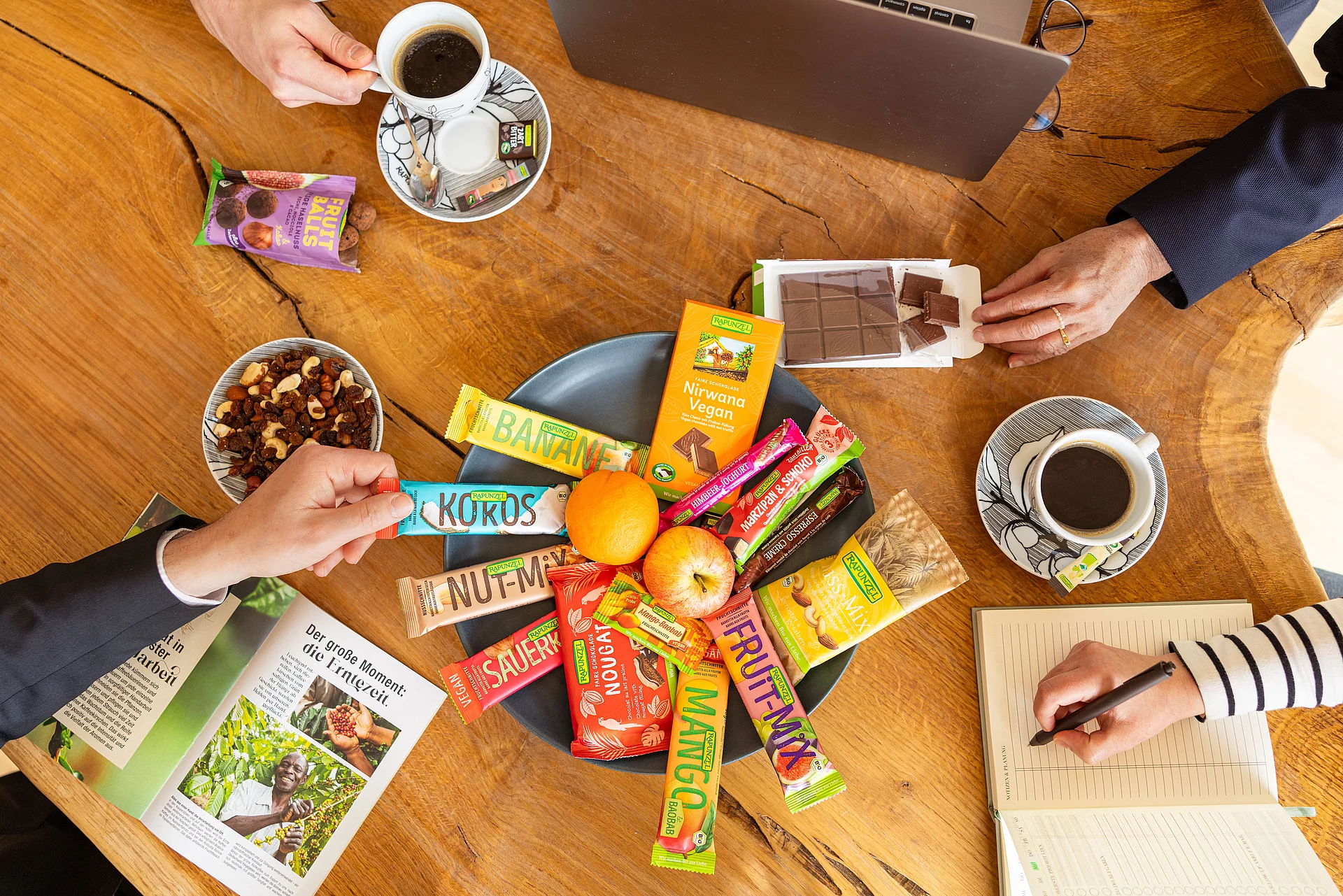 Rapunzel Snackauswahl perfekt für Messen, Tagungen & Co. Verschiedene Rapunzel Snacks in einer Schüssel verteilt auf einem Tisch. Mehrere Menschen greifen nach den Snacks.