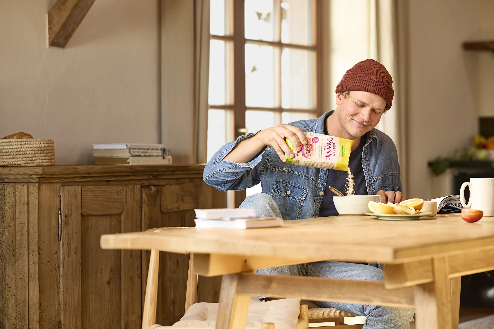 Ein junger Mann sitzt in einer gemütlichen Bauernhofküche und füllt sich Porridge in eine Müslischüssel. Auf dem Tisch liegen Zeitschriften und eine Schale mit.