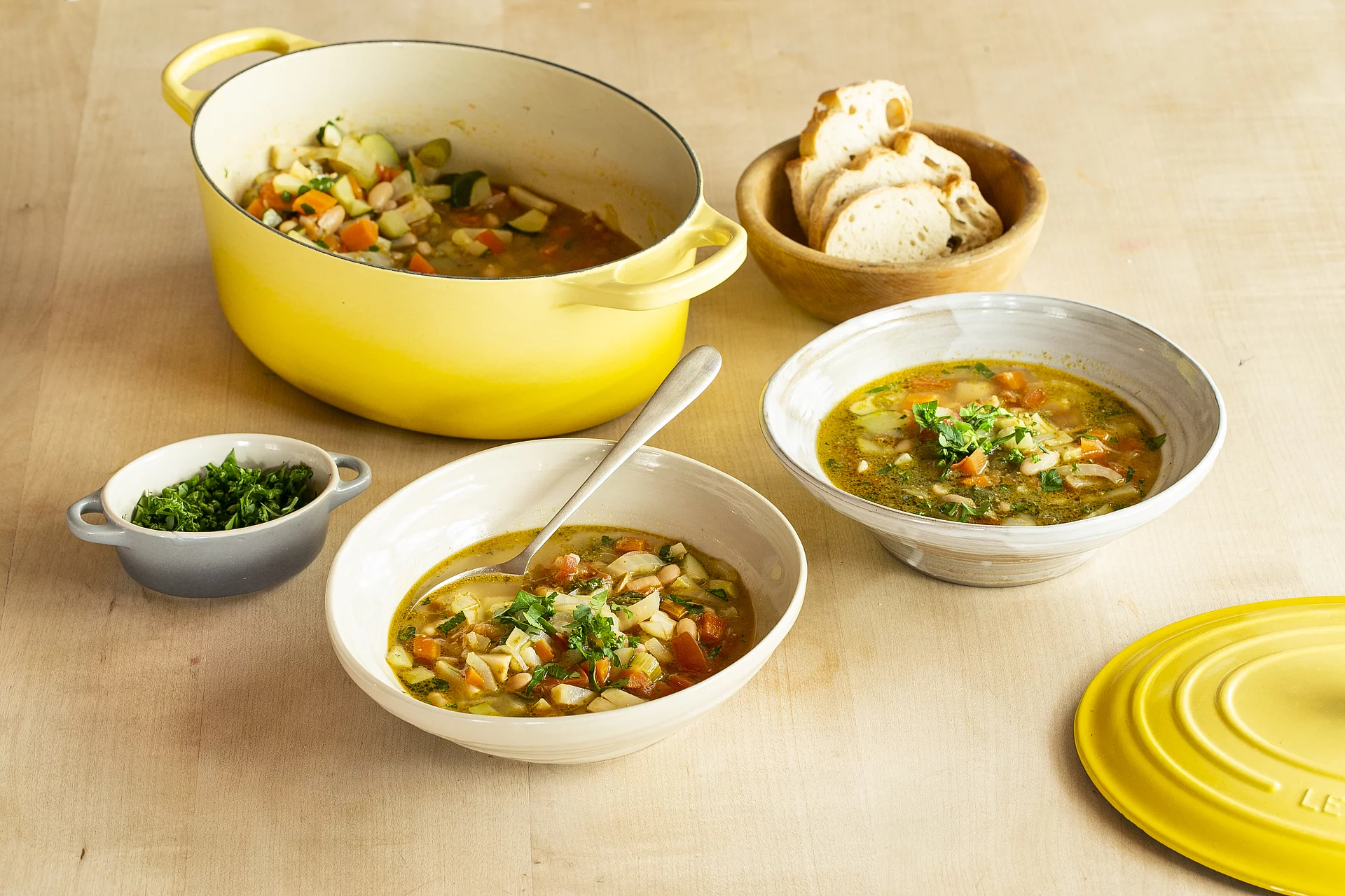 Minestrone - Italienischer Klassiker mit Rapunzel Gemüse-Brühwürfel Original Zwei Teller mit Minestrone neben einem gelben Kochtopf und Schüsseln mit Brot und Kräutern.