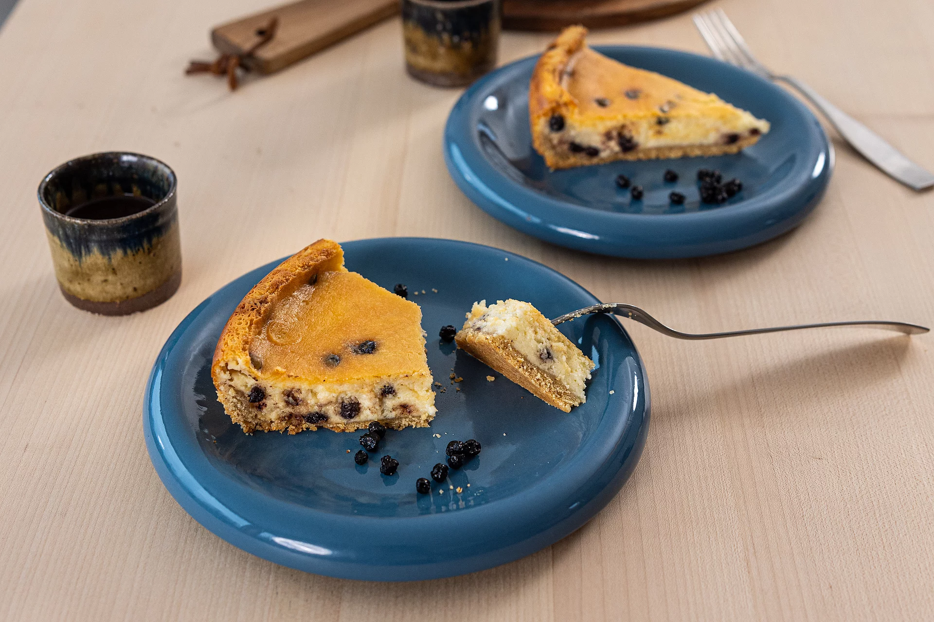 Glutenfreier Käsekuchen mit Erdmandelmehl. Käsekuchenstücke mit Blaubeeren auf zwei Tellern, daneben Becher mit Kaffee.