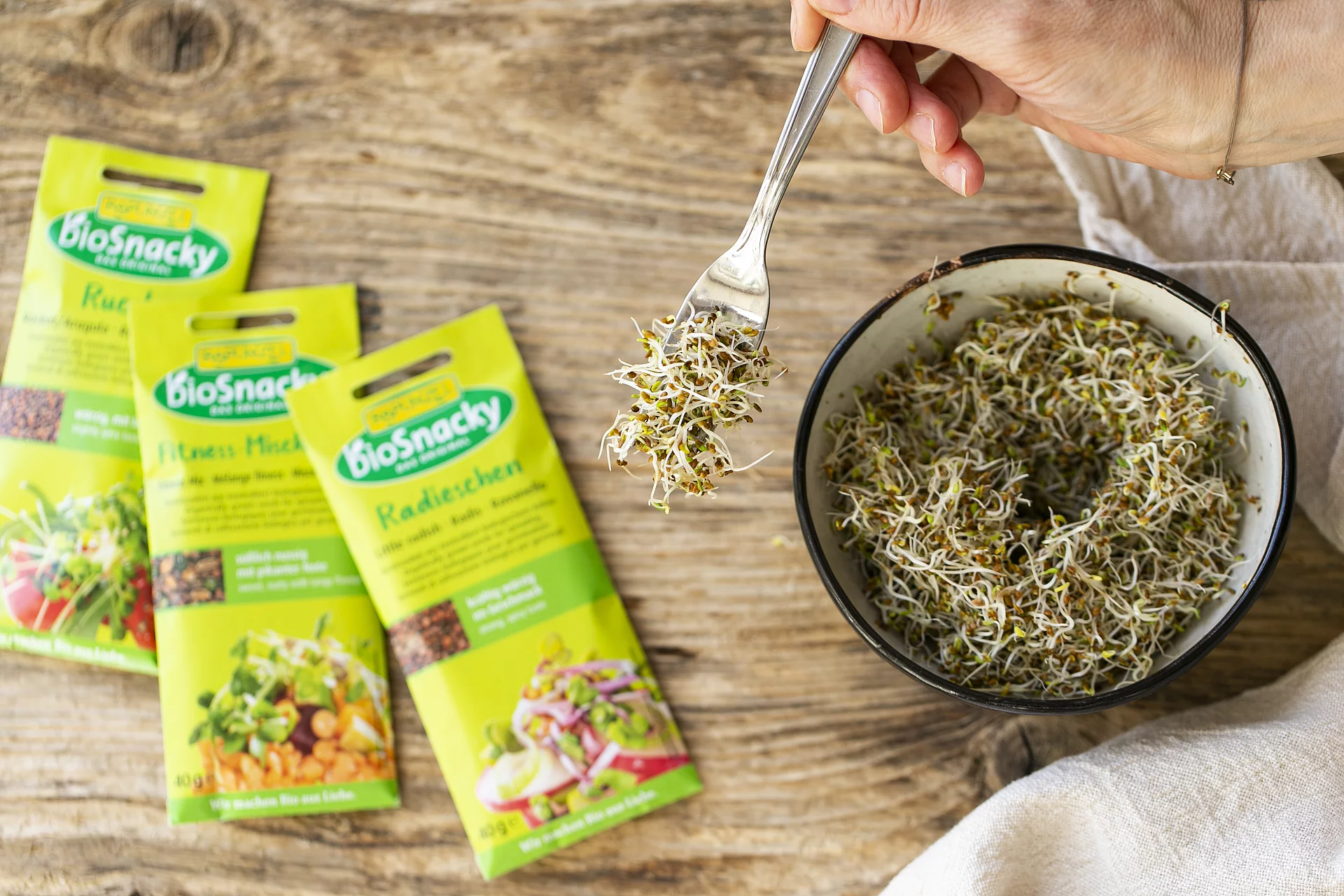 Drei Packungen Keimsaaten von Rapunzel bioSnacky neben Schale mit frischen Sprossen.