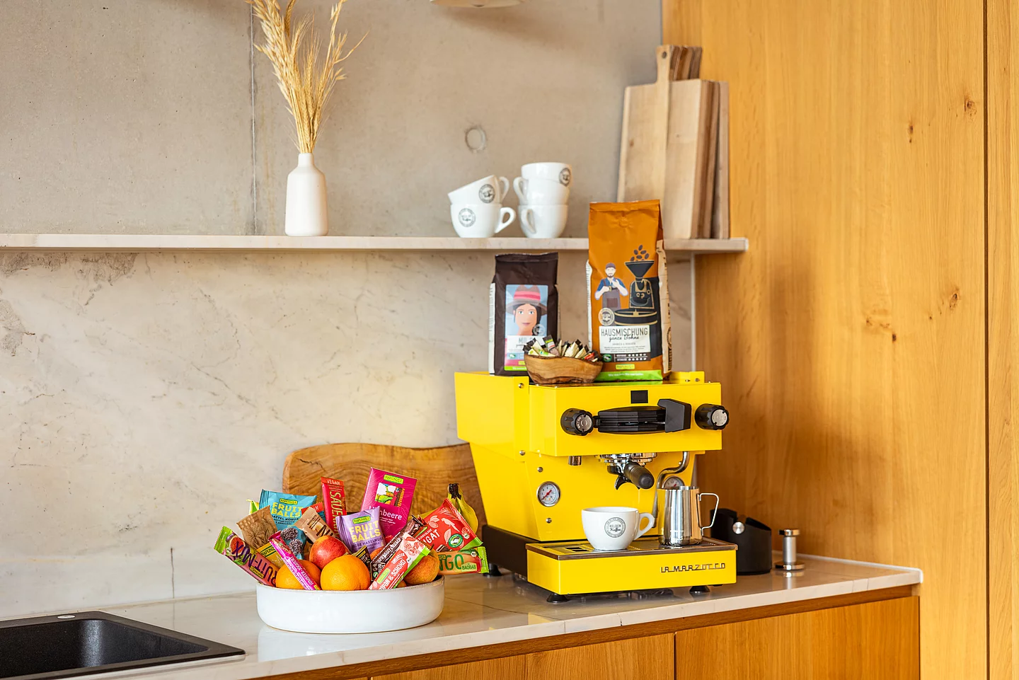 Rapunzel Knabbereien und Snacks - süß und salzig. Eine Schüssel voller Snacks von Rapunzel. Fruchtschnitten, Fruit Balls & Schokolade. Dahinter steht eine gelbe Kaffeemaschine.