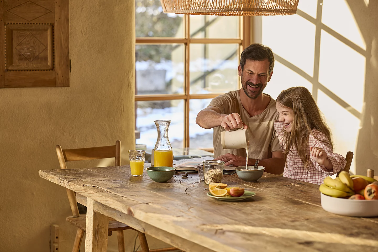 Ein Vater und seine Tochter sitzen gemeinsam am Frühstückstisch. Der Vater schenkt Milch in die Müslischüssel des Kindes und bereitet das Frühstück zu.