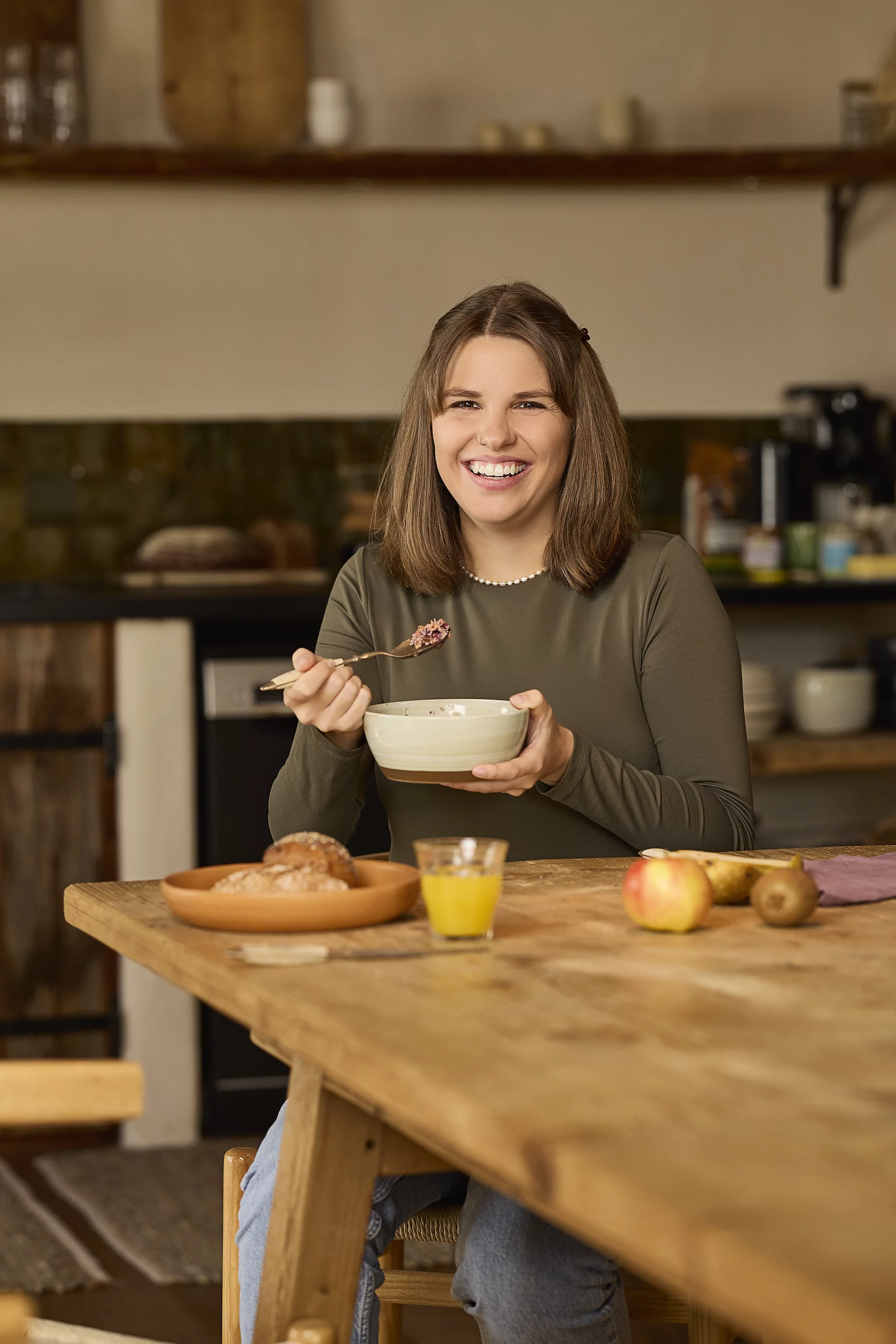 Eine junge Frau sitzt lächelnd am Frühstückstisch und hält eine Schale Beerenmüsli in der Hand. Auf dem Tisch stehen frische Brötchen und ein Glas frisch gepresster Orangensaft.