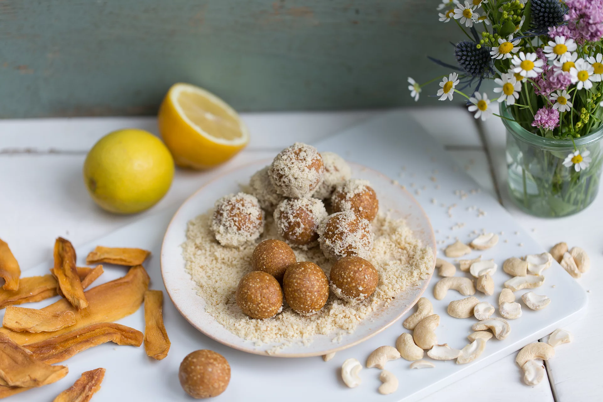 Mango-Cashew-Glückskugeln. Fruchtkugeln aus Erdmandeln auf einem Teller, daneben die Zutaten Mango und Cashewkerne.